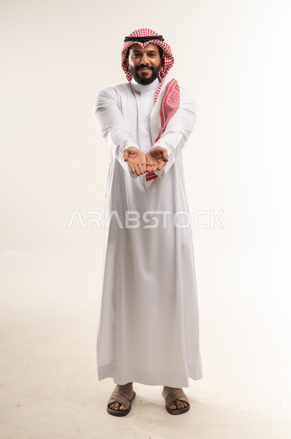 مفهوم التقديم والعرض، بورتريه لرجل عربي خليجي بحريني مبتسم يرتدي الثوب التقليدي والشماغ يفتح كفيه للأعلى بإيماءات حمل عنصر ما, الظر الى الكاميرا بتعابير السعادة والبشاشة، الطول الكامل للجسم، خلفية بيضاء