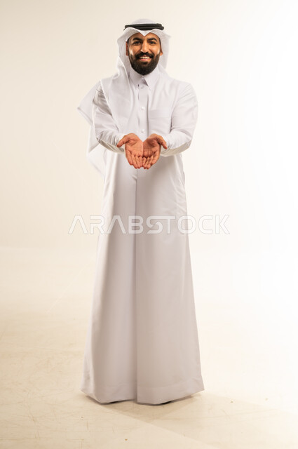 Opening and extending the palms forward, offering hospitality or giving something, elegance, handsomeness and masculinity, the concept of appreciation and respect, portrait of a smiling Qatari Gulf Arab man wearing traditional thobe and ghutra looking at the camera with gestures of happiness and pleasure, full body, white background