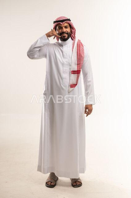 إجراء مكالمة هاتفية بايماءات البشاشة، تعابير الفرح والسرور، انتظار مكالمة وإستخدام الميكروفون، بورتريه لرجل عربي خليجي بحريني مبتسم يرتدي الثوب  التقليدي والشماغ يرفع يده بإيماءات الإتصال، الطول الكامل للجسم, خلفية بيضاء