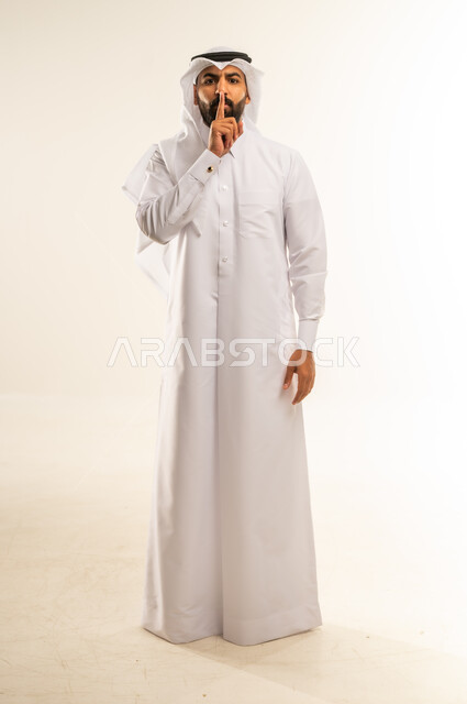 طلب التوقف عن الكلام والصمت، النهي والزجر والانصات، وضع اصبع السبابة أمام الفم بإيماءات الجدية، الأمر بالصمت التام، بورتريه لرجل عربي خليجي قطري يرتدي الثوب التقليدي والغتره ينظر للكاميرا بإيماءات الغضب، الجسم بالطول الكامل، خلفية بيضاء