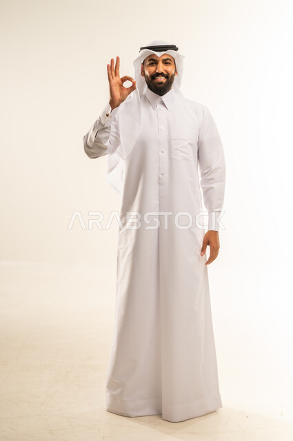التعبير عن الإعجاب والتقدير، رفع اليد بعلامات الجودة والقبول، الامتياز والعلامة الكاملة، الشعور بالرضا والاستحسان، بورتريه لرجل عربي خليجي قطري مبتسم يرتدي الثوب التقليدي والغتره ينظر إلى الكاميرا بإيماءات السعادة والسرور، الجسم بالطول الكامل، خلفية بيضاء