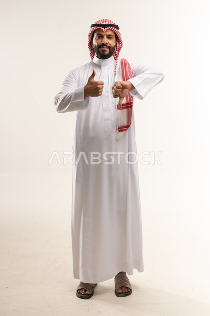 الحيرة والتردد في شيء ما، بورتريه لرجل عربي خليجي بحريني يرتدي الثوب التقليدي والشماغ ينظر الى الكاميرا ويرفع اصبع الابهام للاعلى والاسفل بإيماءات عدم الموافقه والموافقه، تعابير عدم الإعجاب والإعجاب, مفهوم الرفض والقبول، الطول الكامل للجسم, خلفية بيضاء