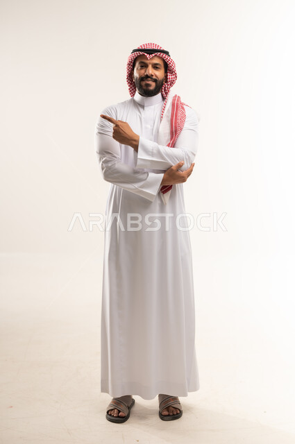 Determine a specific location, indicate the location of an item, portrait of a Bahraini Gulf Arab man wearing a traditional thobe and shemagh pointing with his index finger to a specific target, pointing to the camera with gestures of happiness and pleasure, pointing and explaining something, full body length, white background