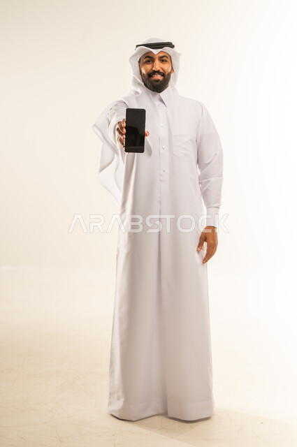 عرض شاشة هاتف محمول سوداء فارغة، دمج التقنيات والتكنولوجيا الحديثة بالحياة اليومية، الإعلانات التسويقية والعروض الترويجية، بورتريه لرجل عربي خليجي قطري مبتسم يرتدي الثوب التقليدي والغتره يمسك الجوال بيده وينظر للكاميرا ببشاشة، الجسم بالطول الكامل، خلفية بيضاء
