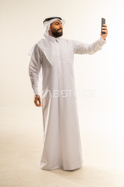 التقاط صور السيلفي باستخدام الهاتف المحمول، دمج التقنيات والتكنولوجيا الحديثة بالحياة اليومية، أخذ اللقطات الشخصية التذكارية، بورتريه لرجل عربي خليجي قطري مبتسم يرتدي الثوب التقليدي والغتره يمسك الجوال بيده وينظر إليه بسعادة، الجسم بالطول الكامل، خلفية بيضاء