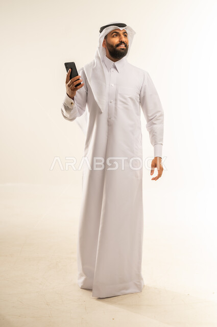 النظر إلى مكان ما بعيد بإيماءات الاندماج، دمج التقنيات والتكنولوجيا بالحياة اليومية، الدردشة والتحدث مع الأصدقاء، تصفح مواقع وتطبيقات الانترنت، بورتريه لرجل عربي خليجي قطري مبتسم يرتدي الثوب التقليدي والغتره يمسك الجوال بيده، الجسم بالطول الكامل، خلفية بيضاء