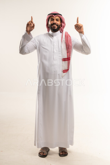 Looking at the camera with gestures of happiness and pleasure, specifying a specific location, pointing and explaining something, portrait of a Bahraini Gulf Arab man wearing a traditional thobe and shemagh raising his index finger up, full body length, blue background