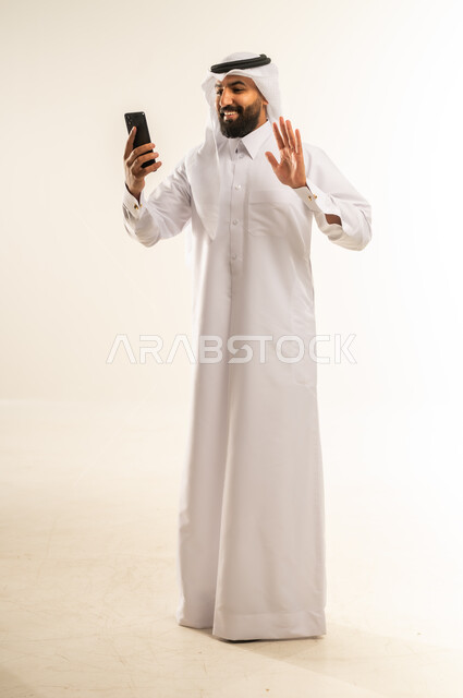 إجراء مكالمات الفيديو عبر الهاتف المحمول، التواصل مع الأهل والأصدقاء، دمج التقنيات الحديثة والتكنولوجيا في الحياة اليومية، بورتريه لرجل عربي خليجي قطري مبتسم يرتدي الثوب التقليدي والغتره يمسك جهاز الجوال بيده بإيماءات السرور، الجسم بالطول الكامل، خلفية بيضاء