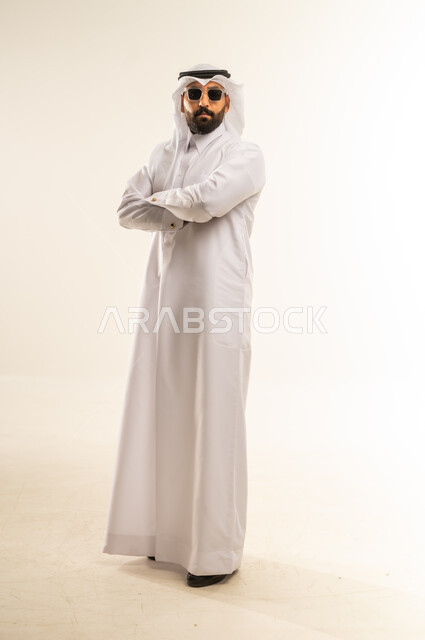 الأناقة والوسامة والاهتمام بالمظهر الخارجي، بورتريه لرجل عربي خليجي قطري مبتسم يرتدي الثوب التقليدي والغتره يقف مكتوف اليدين بإيماءات الثقة بالنفس ويضع نظارة شمسية سوداء، استخدام العدسات الطبية لحماية العين من أشعة الشمس، الجسم بالطول الكامل، خلفية بيضاء