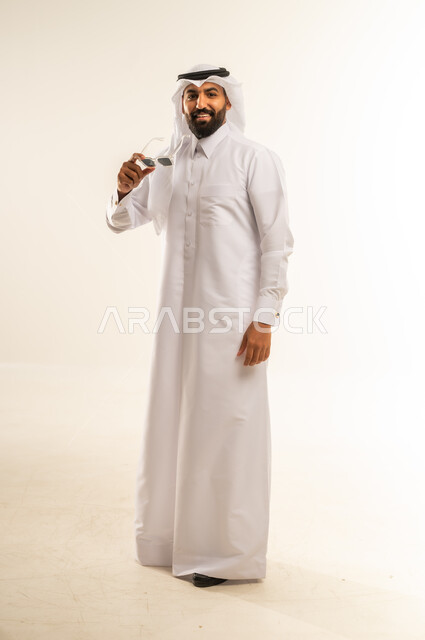 استخدام النظارات الشمسية السوداء لحماية العيون من أشعة الشمس، مفهوم الوسامة والثقة بالنفس، بورتريه لرجل عربي خليجي قطري مبتسم يرتدي الثوب التقليدي والغتره يقف بشكل مستقيم، الرجولة العربية الأصيلة، الاهتمام بالمظهر الخارجي، الجسم بالطول الكامل، خلفية بيضاء