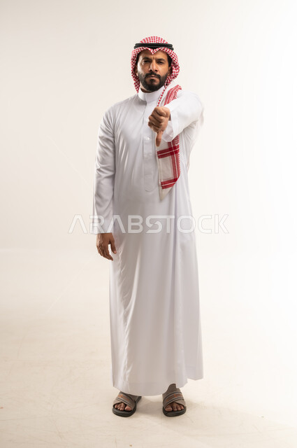 الشعور بالانزعاج من شيء ما، حركات يدين تدل على الرفض وعدم القبول والفشل، بورتريه لرجل عربي خليجي بحريني يرتدي الثوب التقليدي والشماغ يشير بإصبعه الابهام الى الأسفل بإيماءات عدم الاعجاب والتقييم السلبي، الطول الكامل للجسم, خلفية بيضاء