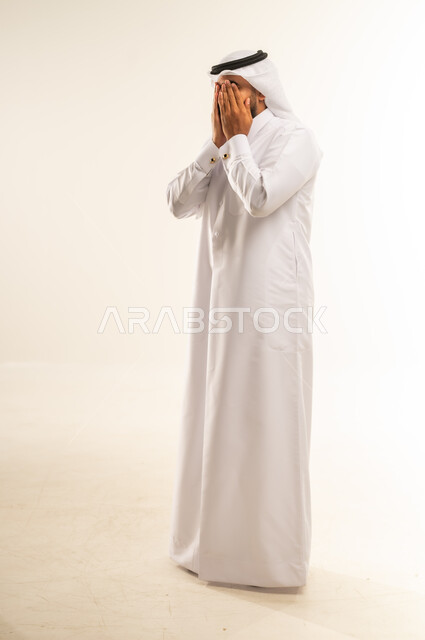 البكاء ومشاعر الحزن، إغماض العينين وعدم الرغبة بالرؤية، الشعور بالخجل والإحراج، التعرض لصدمة وعدم التصديق، بورتريه لرجل عربي خليجي قطري يرتدي الثوب التقليدي والغتره يغطي وجهه بيديه، الجسم بالطول الكامل، خلفية بيضاء