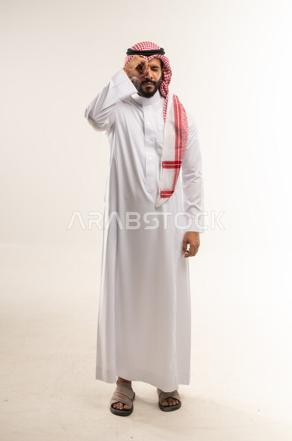 محاولة تقريب الأشياء البعيدة, مفهوم التخطيط و الرؤية المستقبلية، بورتريه لرجل عربي خليجي بحريني يرتدي الثوب التقليدي والشماغ يضع يده على عينيه بشكل عدسة بإيماءات البحث والاستكشاف، التركيز والنظر إلى شيء محدد، الطول الكامل للجسم، خلفية بيضاء