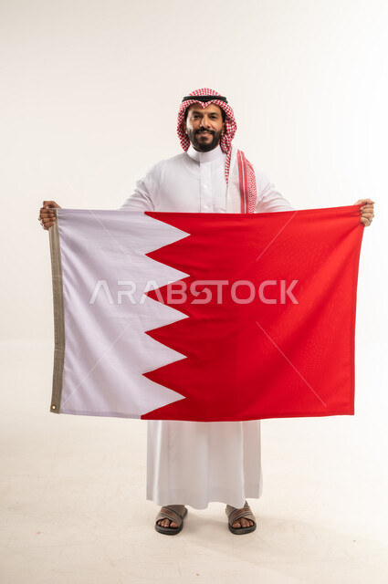 إحياء ذكرى استقلال الدولة 14 أغسطس، الاحتفال باليوم الوطني البحريني 16 ديسمبر، بورتريه لرجل عربي خليجي بحريني يحمل علم مملكة البحرين، رموز وثقافات وطنية, حب الوطن والانتماء إليه، النظر الى الكاميرا بايماءات الفخروالاعتزاز، الطول الكامل للجسم، خلفية بيضاء