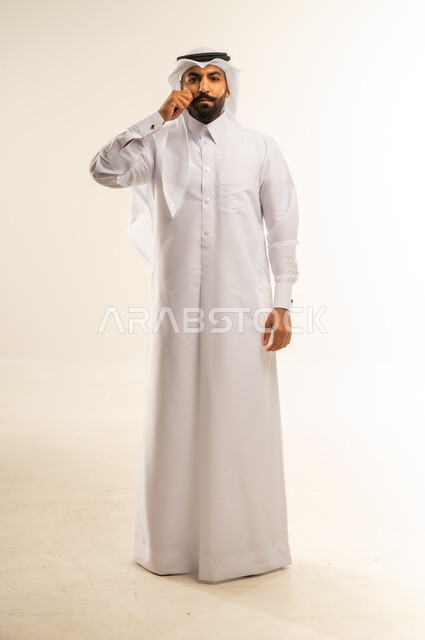 الرجولة العربية الأصيلة، الاهتمام بالمظهر الخارجي، النظر للكاميرا بإيماءات الجدية، مفهوم الوسامة والثقة بالنفس، بورتريه لرجل عربي خليجي قطري يرتدي الثوب التقليدي والغتره يقف بشكل مستقيم ويمسك شاربه، إعطاء الوعود الصادقة، الجسم بالطول الكامل، خلفية بيضاء
