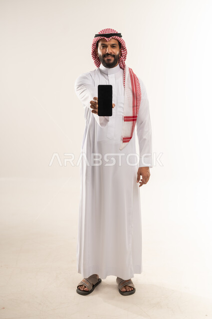 E-marketing and benefiting from mobile services and applications, using modern technologies in daily life, portrait of a Bahraini Gulf Arab man wearing a traditional thobe and shemagh displaying a blank black screen on a mobile phone, full body length, white background