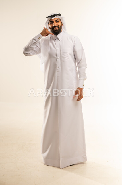 مفهوم تبادل المعلومات، حركة يد تدل على الاتصال، الاهتمام بالمظهر الخارجي والوسامة، بورتريه لرجل عربي خليجي قطري يرتدي الثوب التقليدي والغترة يرفع يده بايماءات الرد على المكالمات الهاتفية، تلقي أخبار سارة عبر الجوال، الجسم بالطول الكامل، خلفية بيضاء
