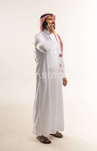 Communication with family and friends, gestures and expressions of happiness and pleasure to hear good news, portrait of a smiling Bahraini Gulf Arab man wearing traditional thobe and shemagh making a phone call via mobile phone, use of modern technologies and devices, full body length, white background