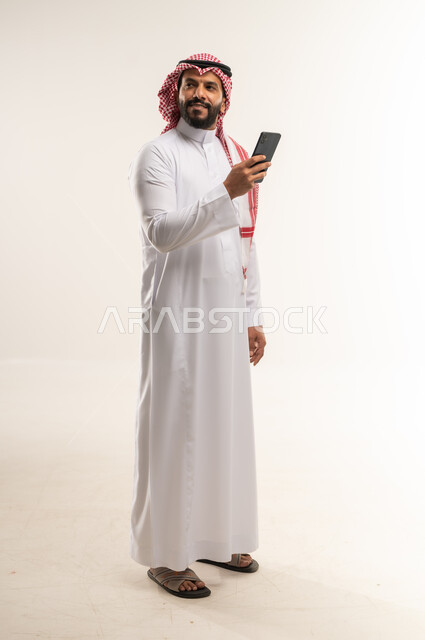 النظر الى شيء ما بايماءات السرور، التواصل مع الافراد عبر الهاتف المحمول، دردشة ومحادثات وتعارف، بورتريه لرجل عربي خليجي بحريني مبتسم يرتدي الثوب التقليدي والشماغ يستعمل الجوال، تصفح الانترنت والتسوق الالكتروني، الطول الكامل للجسم، خلفية بيضاء