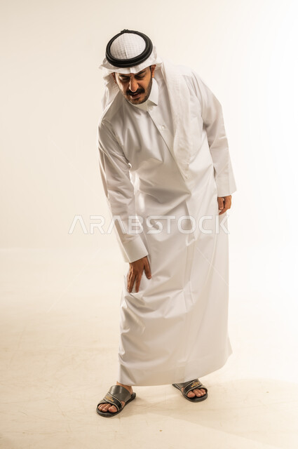 الصعوبة في المشي، التهاب المفاصل والعضلات، آلام العظام والأوتار، الحاجة للذهاب للطبيب والمعاينة، ضرورة الاسترخاء وأخذ قسط من الراحة، بورتريه لرجل عربي خليجي قطري يرتدي الثوب التقليدي والغتره يضع يده على ركبته بإيماءات التألم، الجسم بالطول الكامل، خلفية بيضاء