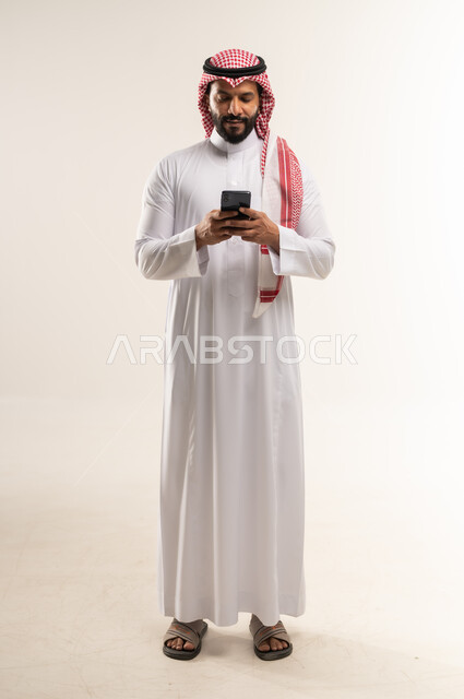 Chat, conversations and dating, portrait of a smiling Bahraini Gulf Arab man wearing traditional thobe and shemagh using a mobile phone, surfing the internet and online shopping, using modern technologies and devices, communicating with individuals via mobile phone, full body length, white background