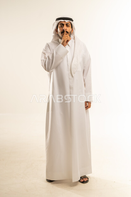 وضع اصبع السبابة أمام الفم بإيماءات الجدية، طلب التوقف عن الكلام والصمت، النهي والزجر والانصات، الأمر بالصمت التام، بورتريه لرجل عربي خليجي قطري يرتدي الثوب التقليدي والغتره ينظر للكاميرا بإيماءات الغضب، الجسم بالطول الكامل، خلفية بيضاء