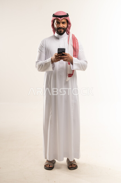 استخدام التقنيات والأجهزة الحديثة، النظر الى الكاميرا ما بايماءات السرور، التواصل مع الافراد عبر الهاتف المحمول، دردشة ومحادثات وتعارف، بورتريه لرجل عربي خليجي بحريني مبتسم يرتدي الثوب التقليدي والشماغ يستعمل الجوال، الطول الكامل للجسم، خلفية بيضاء