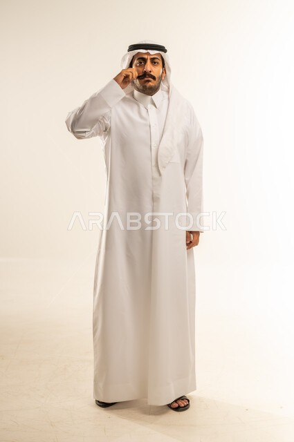 النظر للكاميرا بإيماءات الجدية، الرجولة العربية الأصيلة، الاهتمام بالمظهر الخارجي، مفهوم الوسامة والثقة بالنفس، بورتريه لرجل عربي خليجي قطري مبتسم يرتدي الثوب التقليدي والغتره يقف بشكل مستقيم ويمسك شاربه، إعطاء الوعود الصادقة، الجسم بالطول الكامل، خلفية بيضاء