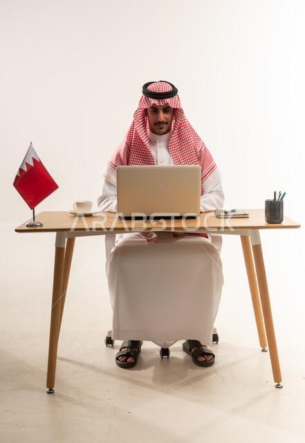 مهن ووظائف مكتبية، دمج التكنولوجيا والتقنيات الحديثة بالحياة العملية، بورتريه لشاب عربي خليجي بحريني يرتدي الثوب التقليدي والشماغ يجلس خلف الطاولة وبجانبه فنجان القهوة وعلم البحرين، العمل على جهاز الحاسب المحمول بايماءات السرور، خلفية بيضاء
