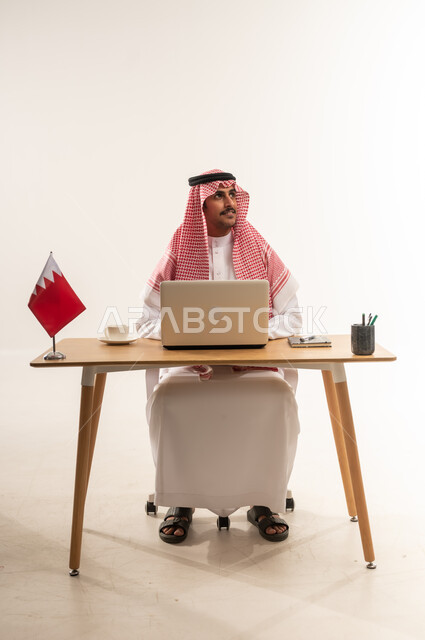 Looking up with gestures of thinking and absent-mindedness, integrating technology and modern techniques into practical life, portrait of a young Arab Gulf Bahraini man wearing traditional thobe and shemagh sitting behind a table with the Bahraini flag next to him, working on a laptop, office jobs and professions, white background