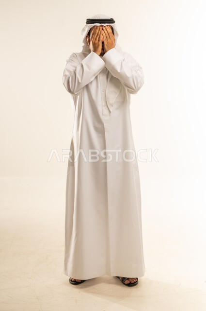 Closing eyes and not wanting to see, feeling shy and embarrassed, crying and feeling sad, being shocked and disbelief, portrait of a Qatari Gulf Arab man wearing a traditional thobe and ghutra covering his face with his hands, full body, white background