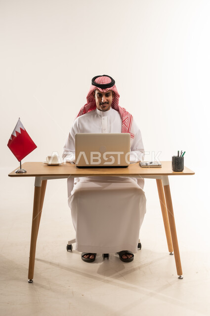 مهن ووظائف مكتبية، دمج التكنولوجيا والتقنيات الحديثة بالحياة العملية، بورتريه لشاب عربي خليجي بحريني يرتدي الثوب التقليدي والشماغ يجلس خلف الطاولة وبجانبه فنجان القهوة وعلم البحرين، العمل على جهاز الحاسب المحمول بايماءات السرور، خلفية بيضاء