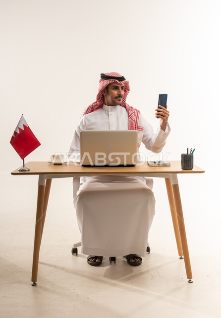 اجراء مقابلات عمل اونلاين عبر الهاتف المحمول، بورتريه لشاب عربي خليجي بحريني يرتدي الثوب التقليدي والشماغ يجلس خلف الطاولة ويعمل على جهاز اللابتوب، استخدام التقنيات الحديثة والتكنولوجيا, مهن ووظائف مكتبية، خلفية بيضاء