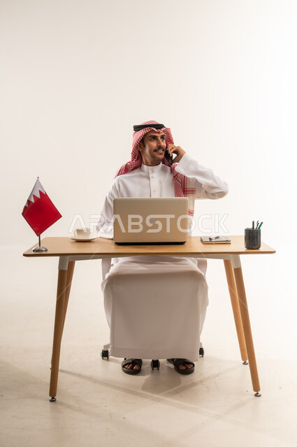 إجراء المكالمات الهاتفية عبر الجوال، استخدام التقنيات الحديثة والتكنولوجيا، دردشة ومحادثات مع الأصدقاء، مهن ووظائف سعودية، بورتريه لشاب عربي خليجي بحريني يرتدي الثوب التقليدي والشماغ يجلس خلف الطاولة يعمل على اللابتوب وبجانبه علم البحرين، خلفية بيضاء