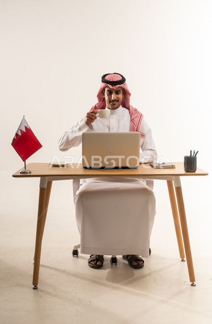 العمل على جهاز الحاسب المحمول بايماءات السرور، مهن ووظائف مكتبية، دمج التكنولوجيا والتقنيات الحديثة بالحياة العملية، بورتريه لشاب عربي خليجي بحريني يرتدي الثوب التقليدي والشماغ يجلس خلف الطاولة يشرب القهوة وبجانبه علم البحرين، خلفية بيضاء