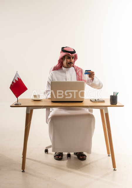 مفهوم التسوق أونلاين، عمليات التبضع والشراء عبر تطبيقات الانترنت، الدفع الالكتروني بالاعتماد على البطاقة البنكية، استخدام التقنيات الحديثة والتكنولوجيا, بورتريه لرجل عربي خليجي بحريني يرتدي الثوب التقليدي والشماغ يعمل على اللابتوب وبجانبه علم البحرين، خلفية بيضاء