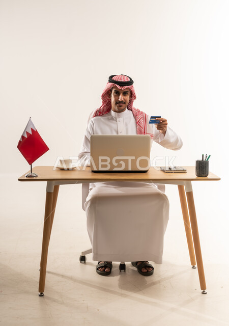 الدفع الالكتروني بالاعتماد على البطاقة البنكية، استخدام التقنيات الحديثة والتكنولوجيا, بورتريه لرجل عربي خليجي بحريني يرتدي الثوب التقليدي والشماغ يعمل على اللابتوب وبجانبه علم البحرين، مفهوم التسوق أونلاين، عمليات التبضع والشراء عبر تطبيقات الانترنت، خلفية بيضاء