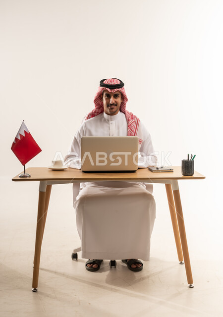 Integrating technology and modern techniques with practical life, portrait of a young Arab Gulf Bahraini man wearing traditional thobe and shemagh sitting behind a table with the Bahraini flag next to him, working on a laptop, office jobs and professions, looking at the camera with gestures of pleasure, white background