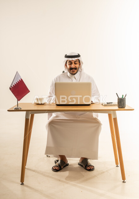 استخدام التقنيات الحديثة في الأعمال الإدارية والمكتبية، التوظيف في المكاتب والشركات القطرية، التركيز والتفاني والعمل الجاد، بورتريه لرجل عربي خليجي قطري يرتدي الثوب التقليدي والغترة يجلس خلف المكتب وبجانبه علم قطر وأمامه الحاسب المحمول، خلفية بيضاء