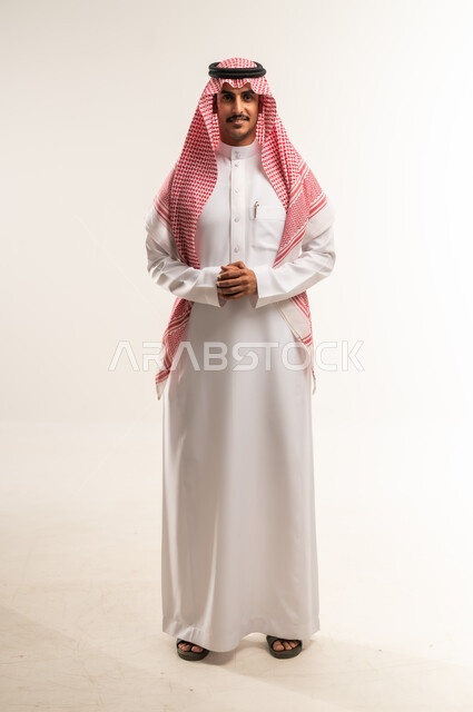تعابير البشاشة والسرور، مفهوم الاناقة والرجولة، الإهتمام بالمظهر الخارجي، بورتريه لرجل عربي خليجي بحريني يرتدي الثوب التقليدي والشماغ يقف بايماءات ايدي متشابكة، النظر إلى الكاميرا بايماءات الثقة بالنفس، الطول الكامل للجسم، خلفية بيضاء