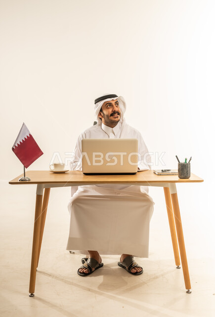 التوظيف في المكاتب والشركات القطرية، التركيز والتفاني والعمل الجاد، استخدام التقنيات الحديثة في الأعمال الإدارية والمكتبية، بورتريه لرجل عربي خليجي قطري يرتدي الثوب التقليدي والغترة يجلس خلف المكتب وبجانبه علم قطر وأمامه الحاسب المحمول، خلفية بيضاء