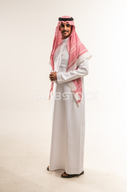 النظر إلى الكاميرا بايماءات الثقة بالنفس، تعابير البشاشة والسرور، مفهوم الاناقة والرجولة، الإهتمام بالمظهر الخارجي، بورتريه لرجل عربي خليجي بحريني يرتدي الثوب التقليدي والشماغ يقف بايماءات ايدي متشابكة، الطول الكامل للجسم، خلفية بيضاء