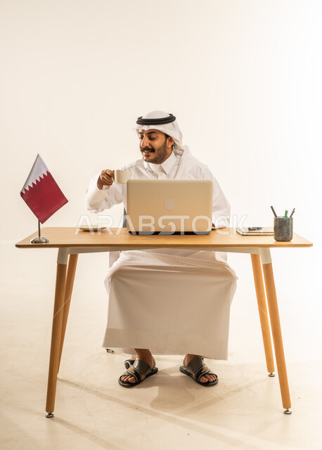 التلذذ بفنجان من القهوة الساخنة، استخدام التقنيات الحديثة بالأعمال الإدارية والمكتبية، التوظيف في المكاتب والشركات القطرية، بورتريه لرجل عربي خليجي قطري يرتدي الثوب التقليدي والغترة يجلس خلف المكتب وبجانبه علم قطر وأمامه الحاسب المحمول، خلفية بيضاء