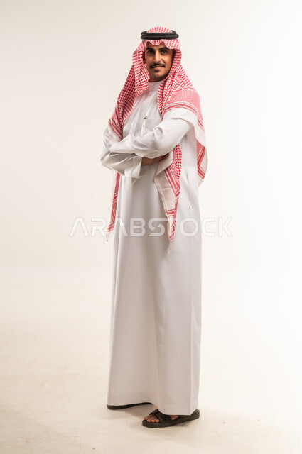 الاهتمام بالأناقة والمظهر الخارجي، النظر إلى الكاميرا بايماءات الجدية والسرور، مفهوم الرجولة والقوة والثقة بالنفس، بورتريه لشاب عربي خليجي بحريني يرتدي الثوب التقليدي والشماغ يقف بأيدي متقاطعة بتعابير الحزم، الطول الكامل للجسم، خلفية بيضاء