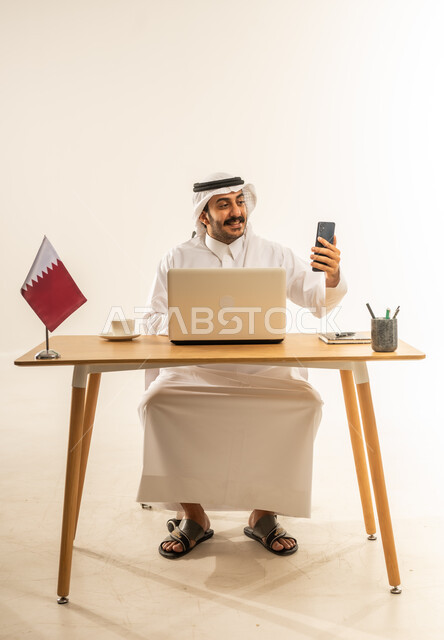 إقامة الاجتماعات أونلاين عبر مكالمات الفيديو، استخدام الهاتف المحمول في العمل، الرد والإجابة على الأسئلة والاستفسارات والمكالمات، بورتريه لرجل عربي خليجي قطري يرتدي الثوب التقليدي والغترة يجلس خلف المكتب أمامه الحاسوب المحمول وعلم قطر، خلفية بيضاء