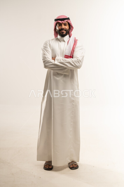 الوقوف بايماءات أيدي متقاطعة بتعابير الحزم والجدية، مفهوم الرجولة والقوة والثقة بالنفس، بورتريه لرجل عربي خليجي بحريني يرتدي الثوب التقليدي والشماغ ينظر الى الكاميرا بتعابير السرور، الاهتمام بالأناقة والمظهر الخارجي، الطول الكامل للجسم، خلفية بيضاء
