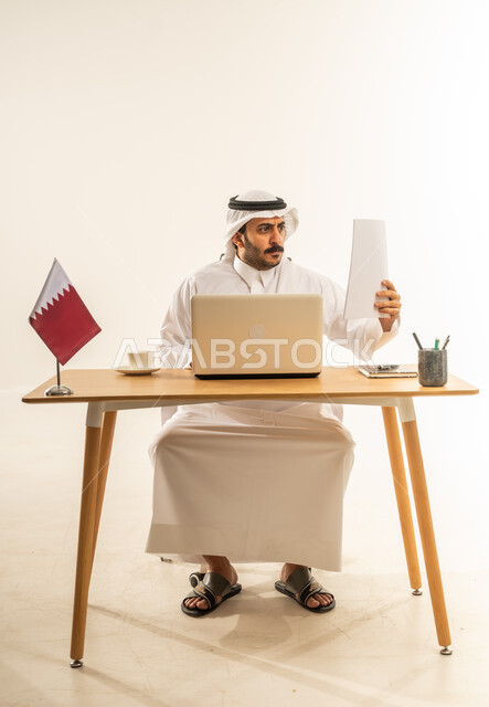 تدقيق الأوراق والملفات الهامة، الاستفادة من التقنيات الحديثة في الأعمال الإدارية والمكتبية، التوظيف في المكاتب والشركات القطرية، بورتريه لرجل عربي خليجي قطري يرتدي الثوب التقليدي والغترة يجلس خلف المكتب وبجانبه علم قطر وأمامه الحاسب المحمول، خلفية بيضاء