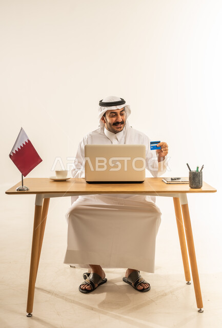 التبضع عن بعد بسهولة ويسر، الشراء عبر تطبيقات ومواقع الانترنت، عملية الدفع الالكتروني بواسطة بطاقة الائتمان، مفهوم التسوق أونلاين، بورتريه لرجل عربي خليجي قطري يرتدي الثوب التقليدي والغترة يجلس خلف المكتب أمامه الحاسوب المحمول وبجانبه علم قطر، خلفية بيضاء