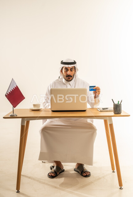 الشراء عبر تطبيقات ومواقع الانترنت، عملية الدفع الالكتروني بواسطة بطاقة الائتمان، مفهوم التسوق أونلاين، التبضع عن بعد بسهولة ويسر، بورتريه لرجل عربي خليجي قطري يرتدي الثوب التقليدي والغترة يجلس خلف المكتب أمامه الحاسوب المحمول وبجانبه علم قطر، خلفية بيضاء