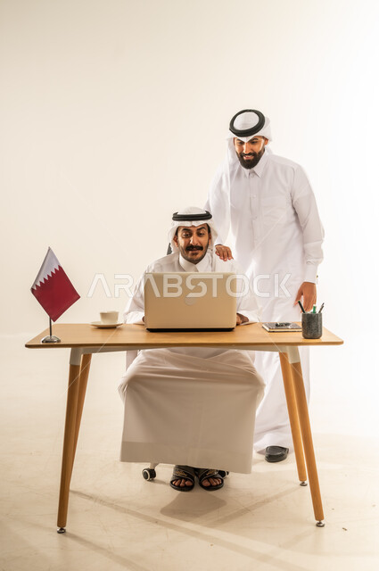 Using laptop in administrative field, concept of teamwork and team spirit, expressing opinion about company strategies and goals, portrait of a Qatari Gulf Arab man wearing traditional thobe and ghutra sitting behind office table with Qatar flag next to him discussing work matters with his colleague, white background